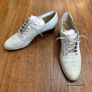 May include: A pair of light grey leather oxford shoes featuring a lace-up closure, a rounded toe, and decorative brogue-style perforations across the front. The shoes have a low stacked heel and include white tissue paper stuffed inside.