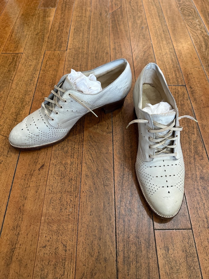 May include: A pair of light grey leather oxford shoes featuring a lace-up closure, a rounded toe, and decorative brogue-style perforations across the front. The shoes have a low stacked heel and include white tissue paper stuffed inside.