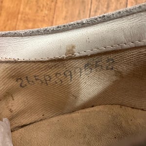 May include: The interior of a shoe featuring a cream-coloured fabric lining and a white leather trim along the top edge. Stamped in black ink on the side of the fabric lining is the alphanumeric code 265P897552, with a small brown stain visible.