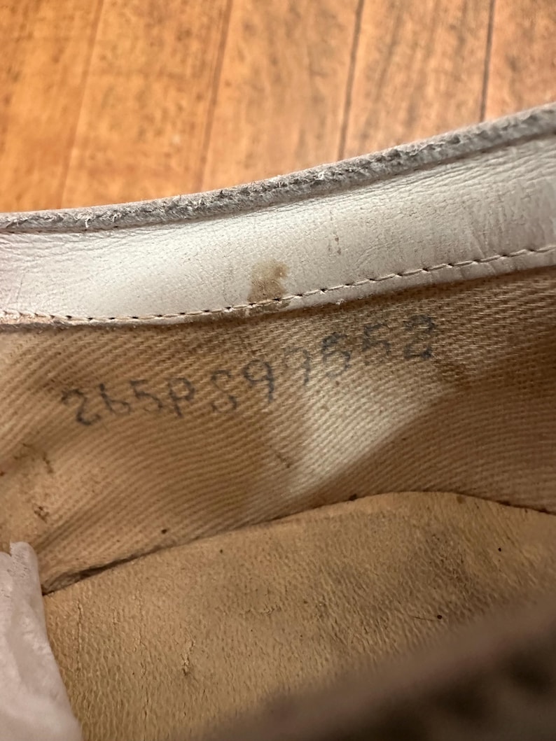 May include: The interior of a shoe featuring a cream-coloured fabric lining and a white leather trim along the top edge. Stamped in black ink on the side of the fabric lining is the alphanumeric code 265P897552, with a small brown stain visible.