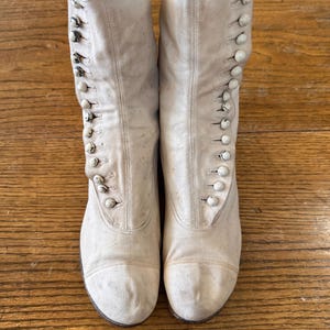 Può includere: Un paio di stivali antichi color crema con bottoni. Gli stivali hanno un design a gamba alta con diversi bottoni sul lato. Gli stivali sono realizzati in un materiale simile alla tela e mostrano segni di usura.