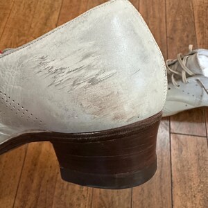 May include: A light grey leather shoe with a low brown stacked heel and visible scuff marks on the side. The shoe features perforated detailing along the side panel and includes light-coloured laces visible on the second shoe in the background.