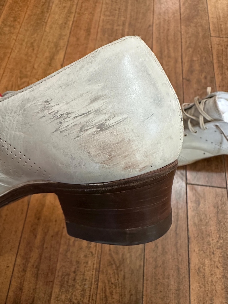 May include: A light grey leather shoe with a low brown stacked heel and visible scuff marks on the side. The shoe features perforated detailing along the side panel and includes light-coloured laces visible on the second shoe in the background.