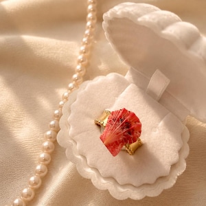 Könnte beinhalten: Ein goldener Ring mit einem rot-weißen Muscheldesign, präsentiert in einer weißen, wellenförmigen Schmuckschatulle. Eine Perlenkette liegt daneben auf einem cremefarbenen Stoff. Der Text "strawberry kiss" steht unten.