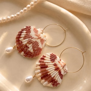 Könnte beinhalten: Ohrringe aus Muscheln mit goldfarbenen Reifen und einer hängenden Perle. Die Muscheln sind weiß mit rotbraunen Streifen. Eine Perlenkette befindet sich im Hintergrund.