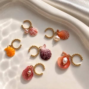 Könnte beinhalten: Ohrringe mit Muscheln und goldfarbenen Reifen. Jeder Ohrring hat eine unterschiedlich farbige Muschel, darunter rosa, orange und lila, und einige haben einen kleinen Perlenakzent. Die Ohrringe sind auf einer weißen Oberfläche angeordnet.