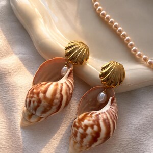 Könnte beinhalten: Ein Paar Ohrhänger mit goldfarbenen, muschelförmigen Steckern, an denen jeweils eine natürliche braun-weiße Spiralschnecke und eine kleine weiße tropfenförmige Perle hängen. Eine passende Kette aus kleinen runden Perlen ist im Hintergrund zu sehen.