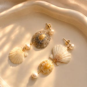Könnte beinhalten: Ohrringe aus Muscheln mit goldfarbenen Akzenten und Kunstperlen. Die Ohrringe zeigen natürliche Muscheln in Weiß, Beige und Braun. Die Ohrringe werden auf einer weißen, glänzenden Oberfläche präsentiert.