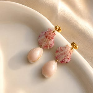 Könnte beinhalten: Ein Paar Ohrhänger mit goldfarbenen Steckern, an denen kleine, fächerförmige rosa-weiße Jakobsmuscheln befestigt sind. Jede Muschel ist mit einer glatten, ovalen, blassrosa Kaurimuschelperle verbunden, was ein hängendes, mehrschichtiges Design ergibt.