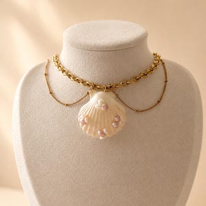 Könnte beinhalten: Eine goldfarbene Kettenhalskette mit einem cremefarbenen Muschelanhänger in der Mitte. Die Muschel ist mit kleinen, runden, schillernden rosa und weißen Perlen verziert. Eine zarte, dünnere Goldkette hängt unter der Hauptgliederkette.