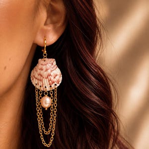Könnte beinhalten: Ein goldfarbener Ohrring mit einer natürlichen Jakobsmuschel mit rosa-weißen radialen Mustern. Die Muschel hängt an einem goldenen Haken und ist mit drei zarten goldenen Ketten sowie einer einzelnen runden, blassrosa Perle darunter verziert.