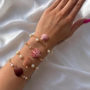 Könnte beinhalten: Vier goldene Kettenarmbänder mit Muscheln und kleinen weißen Perlen. Die Muscheln sind in verschiedenen Rosa- und Brauntönen gehalten. Die Armbänder werden auf einem Arm vor weißem Hintergrund gezeigt.