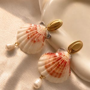 Könnte beinhalten: Muschelohrringe. Jeder Ohrring hat eine natürliche Muschel mit roten und weißen Mustern, einen goldfarbenen ovalen Stecker und eine baumelnde weiße Perle. Die Ohrringe sind für durchbohrte Ohren konzipiert.