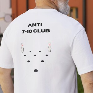 Könnte beinhalten: Weißes T-Shirt mit dem Text "ANTI 7-10 CLUB" und einer Bowling-Pin-Grafik. Das Shirt hat kurze Ärmel und einen Rundhalsausschnitt. Die Grafik zeigt zwei Bowling-Pins und mehrere schwarze Punkte. Das Shirt wird von einer Person getragen.