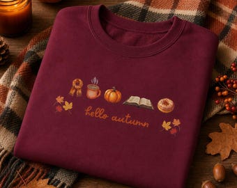 May include: A maroon crewneck sweatshirt featuring a graphic print of a scarf, a mug, a pumpkin, an open book, and a doughnut, with the text "hello autumn" in orange cursive script, accented by small orange and yellow autumn leaf illustrations.