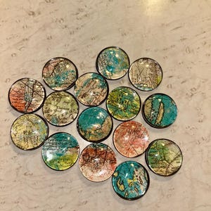 Puede incluir: Una colección de quince botones redondos con tapa de cristal, cada uno con un diseño de mapa vintage. Los mapas presentan una variedad de colores, incluyendo azul, verde, naranja y amarillo, con un borde negro. Los botones están dispuestos sobre una superficie clara.