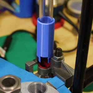 May include: A reloading tool setup featuring a blue plastic funnel and clear tube. The funnel is positioned over a metal component with threaded openings. The setup is on a blue surface, with other tools and components visible in the background.