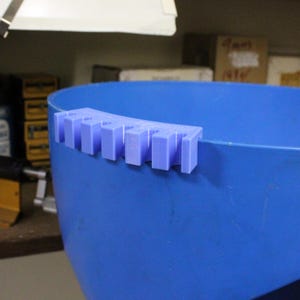 May include: A light purple, 3D-printed tool holder attached to the rim of a blue container. The holder has multiple rectangular slots designed to hold tools. The container is a large, blue, plastic tub.
