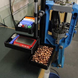 May include: A reloading press with a blue frame and black trays. The trays hold various reloading supplies, including bullets, scales, and other components. The image shows a close-up of the reloading equipment.