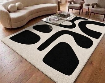 Puede incluir: Una alfombra rectangular con una base de color crema y grandes formas orgánicas abstractas de color negro. La alfombra tiene una superficie de pelo texturizado y está colocada sobre un suelo de madera en una habitación con un sofá curvo.