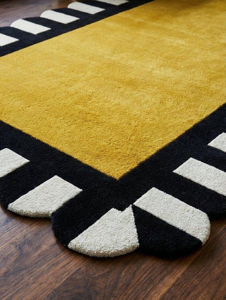 May include: A rectangular area rug featuring a large mustard yellow center panel surrounded by a thick black border. The border is decorated with a repeating pattern of white rectangular shapes along the scalloped edges of the rug.