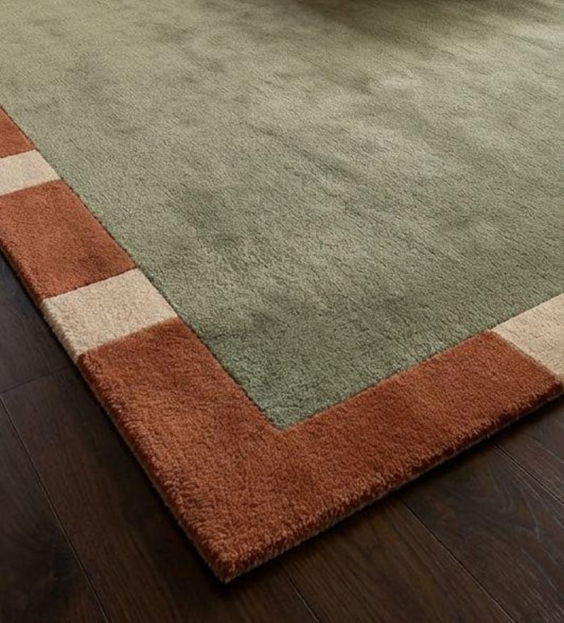 May include: A rectangular area rug with a sage green center and a border of alternating rust and beige rectangles. The rug is made of a soft, plush material, and is placed on a dark wood floor.