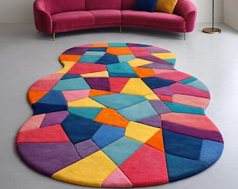 May include: A colorful, abstract rug with a unique shape. The rug features a patchwork design of various geometric shapes in shades of red, blue, yellow, orange, and purple. A magenta sofa with gold legs and a blue and yellow pillow are in the background.
