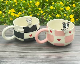 May include: Two ceramic mugs featuring a 3D dog figurine peering over the rim. The left mug has a black and white checkerboard pattern with small red hearts. The right mug has a pink and white checkerboard pattern with small red hearts.