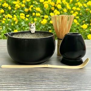 May include: A black ceramic matcha bowl with a small cat figurine on the rim, a bamboo whisk, a black whisk holder, and a bamboo scoop. The items are arranged on a wooden surface with a backdrop of yellow flowers.