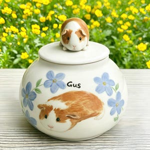 Pode incluir: Um pote de cerâmica branca com uma tampa que apresenta uma figura esculpida de porquinho-da-índia castanho e branco. O pote é decorado com flores azuis pintadas à mão e uma ilustração de um porquinho-da-índia com o nome Gus escrito.