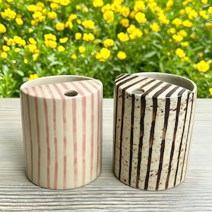 May include: Two ceramic vases with a unique design. One vase is cream-colored with pink vertical stripes, and the other is cream-colored with brown vertical stripes. Both vases have a single hole at the top and a curved top edge.