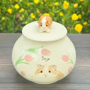 Pode incluir: Um pote de cerâmica creme com tampa, com um desenho de cobaia pintado à mão e a palavra "Joy". A tampa é encimada por uma pequena estatueta de cobaia. Acentos de tulipas cor-de-rosa decoram o pote.