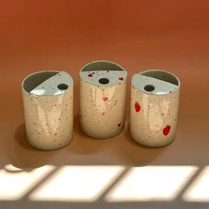 May include: Three speckled ceramic vases with a cylindrical shape and a slanted top opening. The vases are off-white with colorful flecks and small red heart and strawberry designs. The background is a solid orange color.