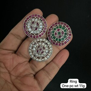 Può includere: Tre anelli argentati con disegni circolari. Ogni anello presenta un motivo centrale di pietre preziose, con pietre rosse e bianche in due anelli e pietre rosse e verdi nel terzo. Gli anelli sono esposti su uno sfondo scuro.