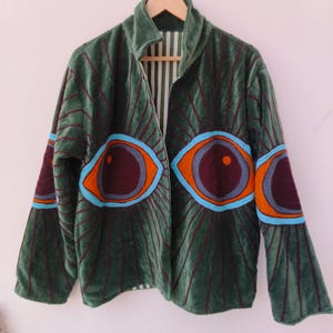 Peut inclure: Veste en velours vert au design unique. La veste présente de grands motifs d'yeux brodés en orange, bleu et bordeaux. Des lignes verticales bordeaux créent un motif texturé. Col montant et doublure rayée.