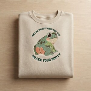 Op de afbeelding: Een beige sweater met ronde hals en een geborduurde groene kikker met donkere stippen en een perzikkleurig achterwerk. De tekst "WHY BE MOODY WHEN YOU CAN SHAKE YOUR BOOTY" is in donkergroene letters in een cirkelvorm rond de kikker geborduurd.