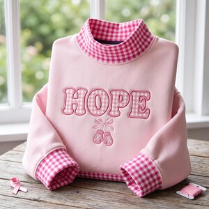 Op de afbeelding: Een lichtroze sweatshirt met een opstaande kraag en mouwboorden in een roze-wit vichy-ruitpatroon. Het woord HOPE is in het midden geborduurd in bijpassende vichy-stof, met daaronder een paar kleine roze bokshandschoenen geborduurd.