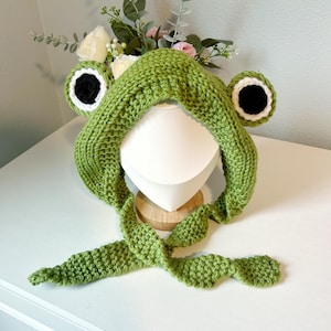 May include: A green knitted frog hat with large, round eyes featuring black pupils and white and black accents. The hat has a hood and long, leaf-shaped ties. The hat is displayed on a white mannequin head.