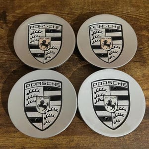 May include: Four silver Porsche wheel center caps. Each cap features the Porsche logo, a black and gold shield with a prancing horse, and the word "Porsche" above. The word "Stuttgart" is also visible.