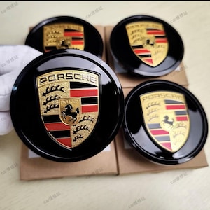 May include: Four black Porsche wheel center caps. Each cap features the Porsche emblem in gold, red, and black, with the word "PORSCHE" above and "STUTTGART" below. The caps are round and have a glossy finish.