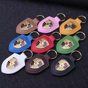 May include: A collection of leather keychains in various colors, including brown, pink, yellow, blue, red, green, white, dark brown, and black. Each keychain features a gold-colored emblem and a metal keyring.