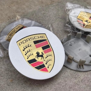 May include: Several silver and white Porsche wheel center caps. Each cap features the Porsche crest in gold, black, red, and yellow, with the word "PORSCHE" above. The caps are new and in their packaging.