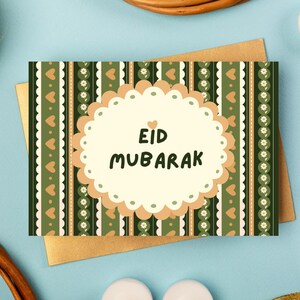 Może przedstawiać: Kartka okolicznościowa z napisem "Eid Mubarak" czarnym pismem na kremowym owalu. Karta ma zielono-beżowo-biały wzór w paski z sercami i kwiatami. Karta leży na złotej kopercie.