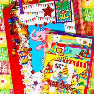 May include: A colorful scrapbook or journal featuring a pink elephant illustration, a clown graphic with the text Clown'n Code, party bunting, and star accents. The cover is decorated with a purple clip, multi-colored beads, and white flower appliques.