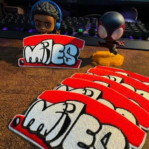 May include: Embroidered patches in red and white, featuring the name "MILES" in a graffiti style. The image also shows two character figurines, one with headphones and the other in a Spider-Man costume.