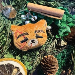 May include: A plush orange cat face with black embroidered features and red scar markings. The cat is surrounded by greenery, a cinnamon stick, a pine cone, and a dried citrus slice. The image has a rustic, natural theme.