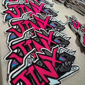 May include: Embroidered patches displaying the word "STINK" in bright pink graffiti lettering. The patches feature a monster design with black and grey details, outlined in light blue. A stack of similar patches is visible.