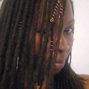 以下が含まれることがあります： 銅色のヘアスパイラルで飾られた、濃い茶色のドレッドヘアの人物。髪は顔を部分的に隠し、ノーズリングが見えます。背景はプレーンな白い壁です。