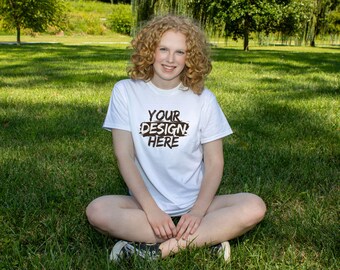 Puede incluir: Una camiseta blanca con el texto "YOUR DESIGN HERE" en fuente negra desgastada. La persona está sentada con las piernas cruzadas sobre un césped. La camiseta es de cuello redondo y manga corta.