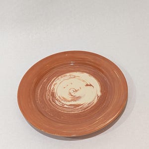 May include: A round ceramic plate featuring a terracotta-colored rim and a contrasting cream-colored center. The surface displays visible brushstroke textures in a circular pattern, creating a swirling effect that blends the two colors together.